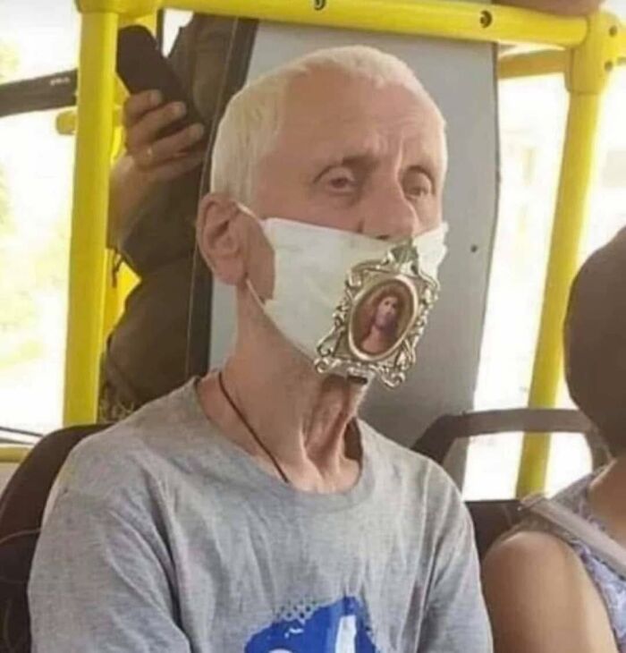 Elderly man on a bus wearing a mask decorated with a small framed religious image, blending occult and unusual humor.