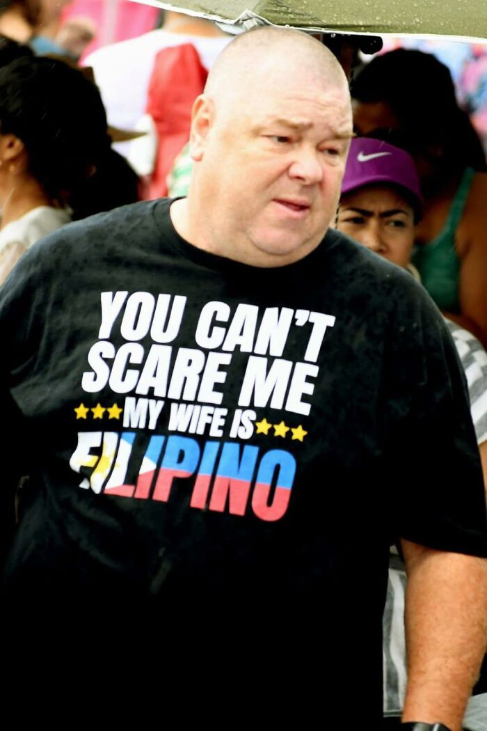 Man wearing a black shirt with a humorous message, surrounded by people, in a candid street scene from pleasures of the occult.