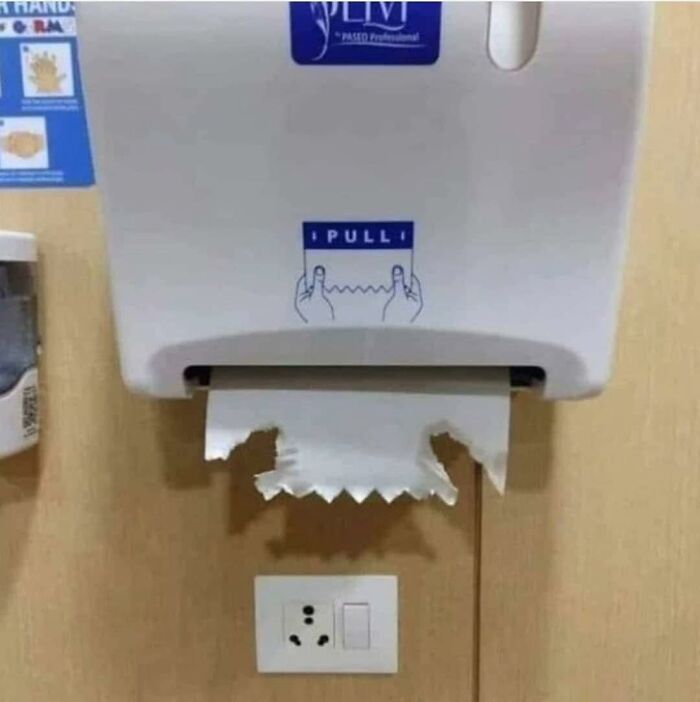 Paper towel dispenser with torn paper towels positioned directly above an electrical outlet in a humorous out of context setting.