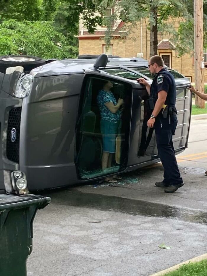 Police officer talking to woman trapped inside overturned car in a bizarre pleasures of the occult moment on a residential street.