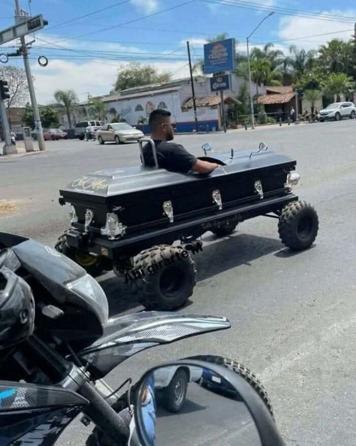 Man driving a coffin modified as an off-road vehicle on a city street, a strange and humorous pleasures of the occult image.