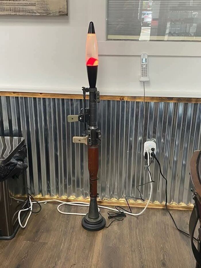 Lava lamp shaped like a rocket launcher standing on a wooden floor in an indoor office with corrugated metal walls and cords.