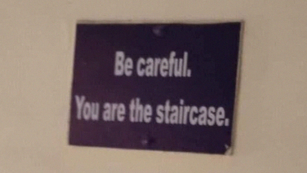 Sign on wall with blurred white text warning to be careful you are the staircase, in an out of context occult humor setting.