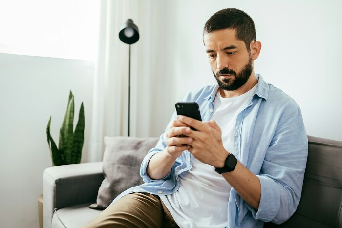 Man sitting on a sofa looking at his phone, reflecting on feelings of insecurity and suspecting his girlfriend&rsquo;s ex&rsquo;s attention.