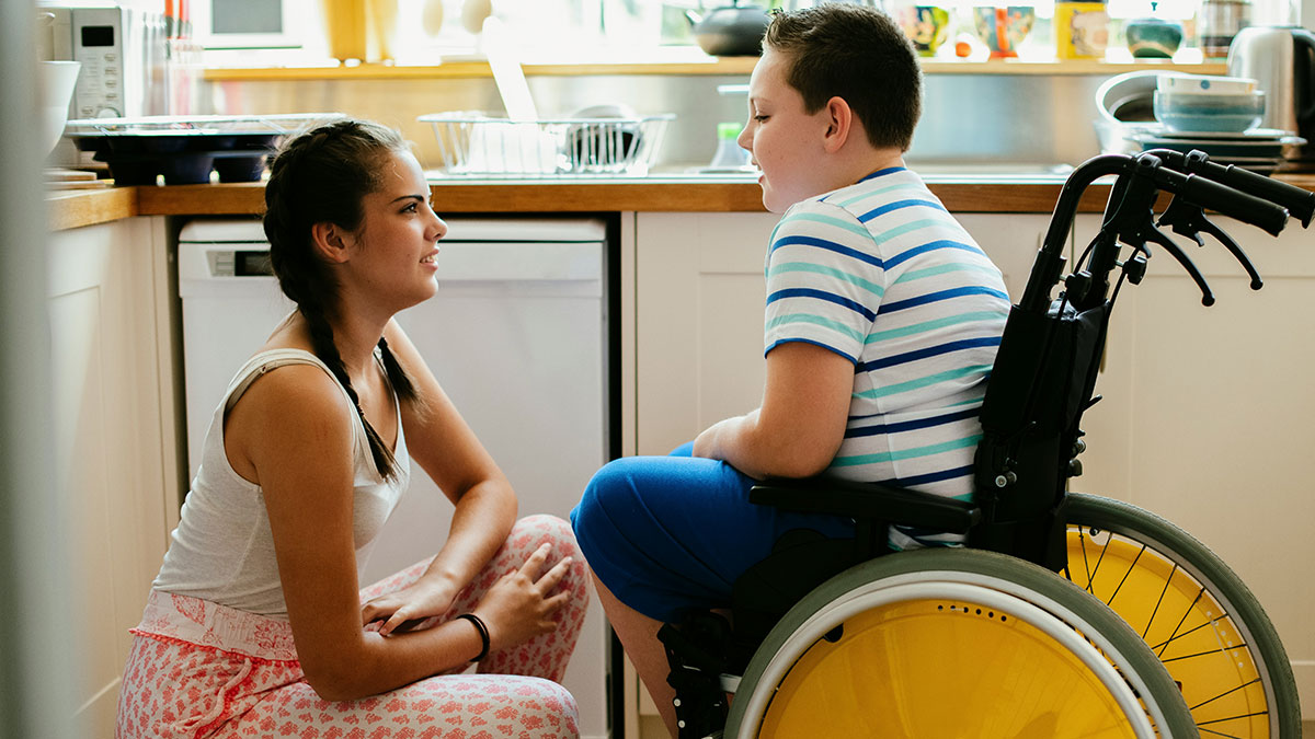 Young babysitter talking with a disabled child in a wheelchair inside a kitchen, showing care and attention