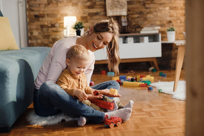 Young child playing with toys on the floor with smiling bio mom in cozy living room, highlighting kids prefer bio mom.