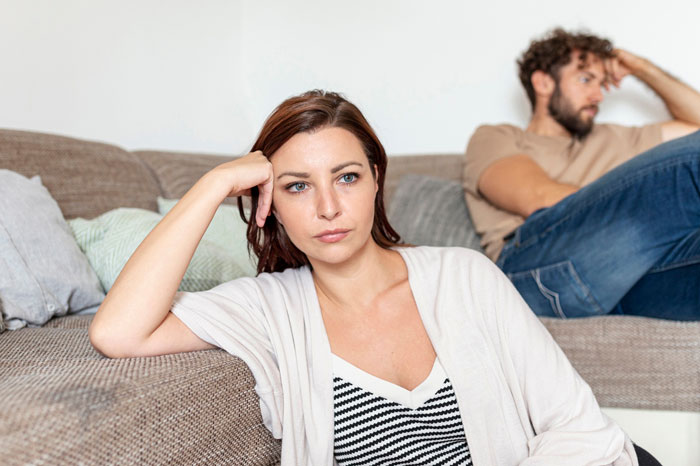 Frustrated wife sitting on couch with upset manchild playing games in background, tense family moment over lost dog. Frustrated wife sitting on couch with upset manchild playing games in background, tense family moment over lost dog.