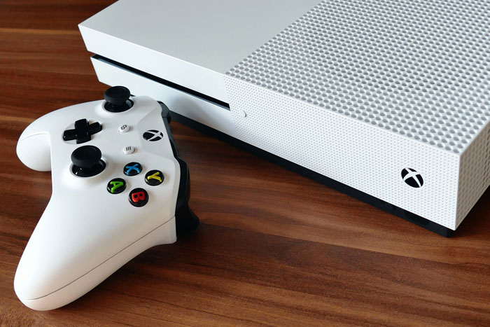 White Xbox console and controller resting on a wooden table, highlighting gaming console and controller details. White Xbox console and controller resting on a wooden table, highlighting gaming console and controller details.
