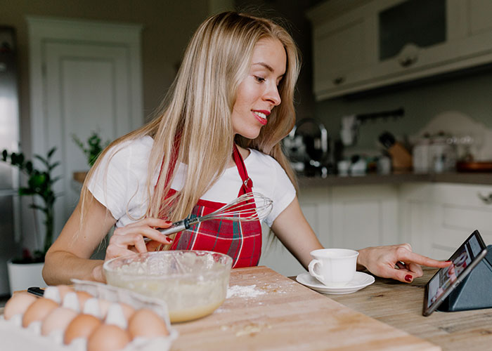 Woman in a kitchen wearing a red apron, holding a whisk and interacting with a tablet while baking stopped for colleagues. Woman in a kitchen wearing a red apron, holding a whisk and interacting with a tablet while baking stopped for colleagues.