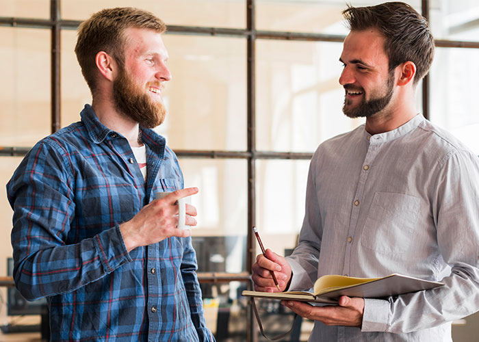 Two male colleagues smiling and chatting in a modern office discussing work and making things awkward at work.