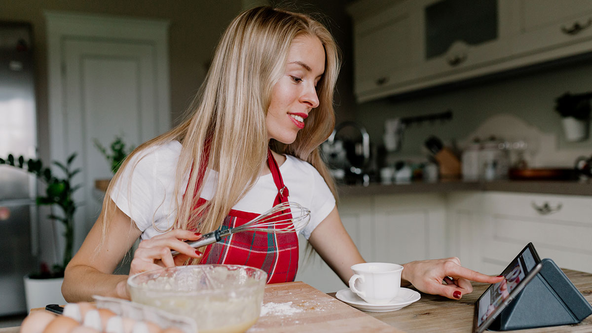 Woman in kitchen wearing red apron, holding whisk and checking tablet while preparing baking ingredients nearby.