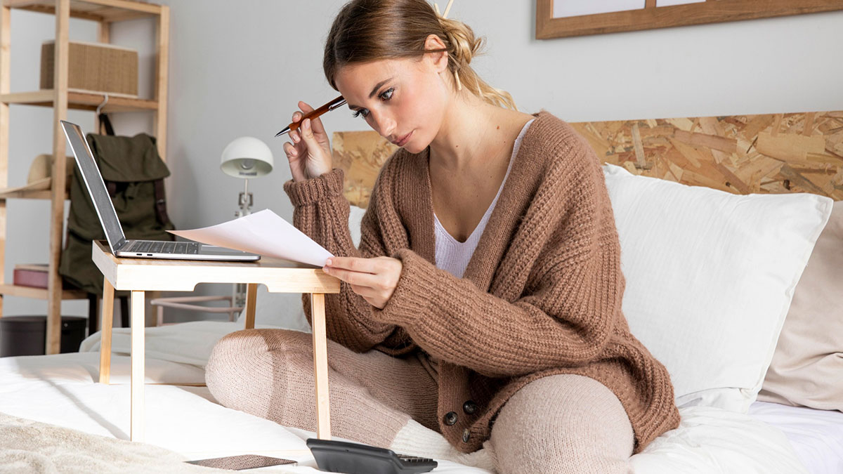Young woman in cozy sweater using laptop and calculator on bed while reviewing expenses for birthday trip planning