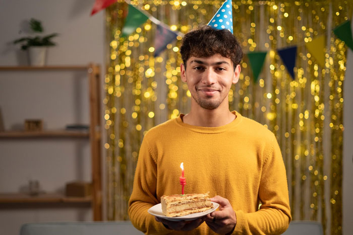 Young man in a party hat holding a birthday cake slice with a candle during a birthday trip celebration surprise