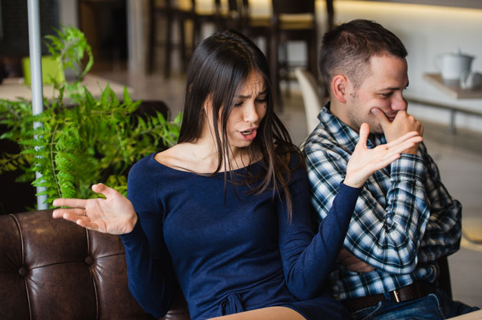 Couple arguing on a couch with girlfriends revealing she covered expenses for birthday trip, friends shocked by the news.