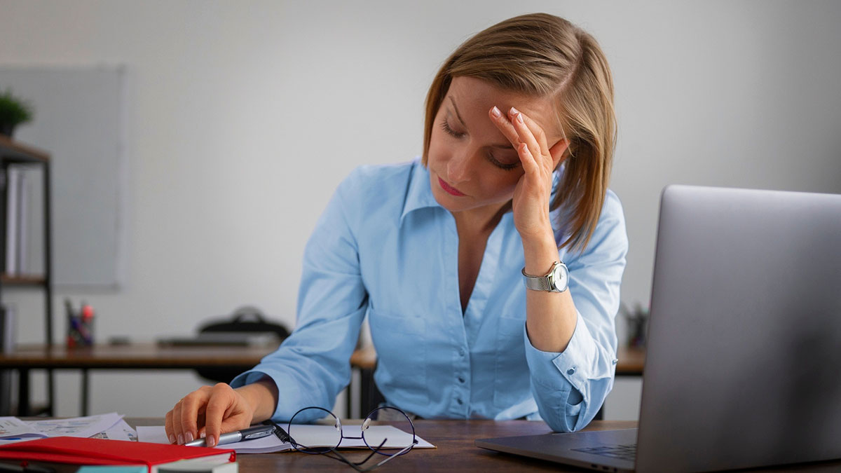 Woman in blue shirt looking stressed at desk, frustrated by ex-coworker taking credit for her work and promotion loss