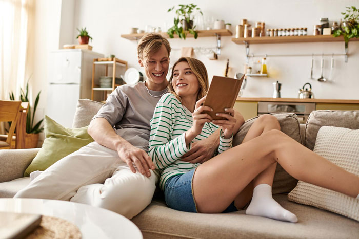 Young couple sitting close on a couch, smiling and reading a book, depicting a relaxed relationship break every year moment. Young couple sitting close on a couch, smiling and reading a book, depicting a relaxed relationship break every year moment.