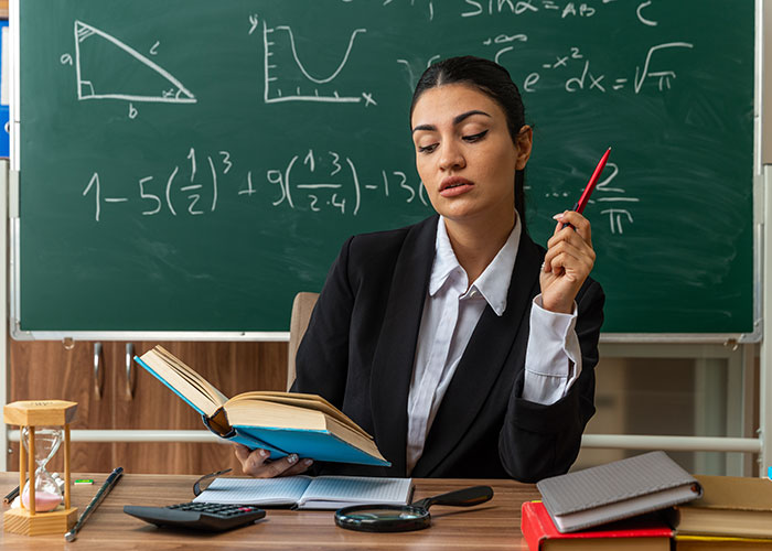 Woman in black suit holding a book and pen, sitting in front of a chalkboard with math formulas, symbolizing rich boyfriend demands split rent. Woman in black suit holding a book and pen, sitting in front of a chalkboard with math formulas, symbolizing rich boyfriend demands split rent.