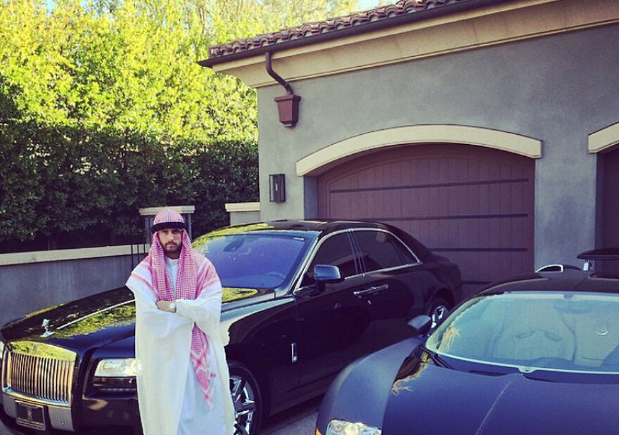 Man in a traditional Middle Eastern costume standing between luxury cars, showing a Halloween costume lacking common sense.