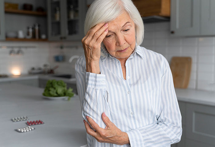Older woman in striped shirt looking worried and stressed while standing in a kitchen near medication pills. Older woman in striped shirt looking worried and stressed while standing in a kitchen near medication pills.