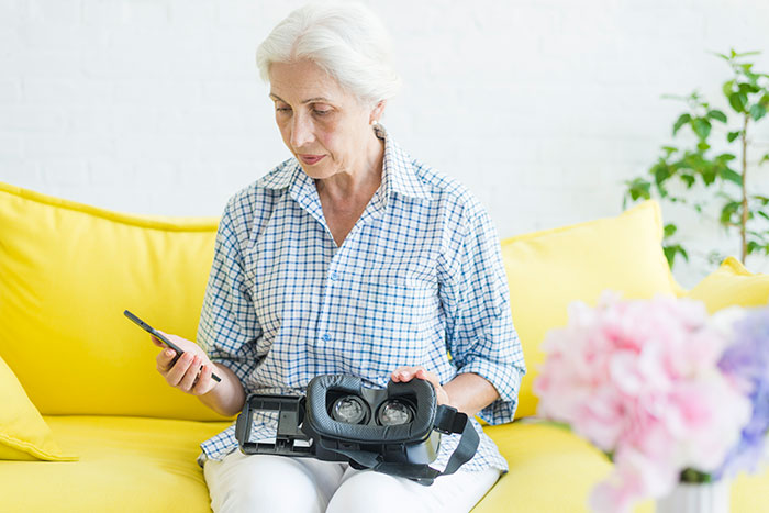 Elderly woman sitting on a yellow couch, looking concerned while holding a phone and a virtual reality headset.