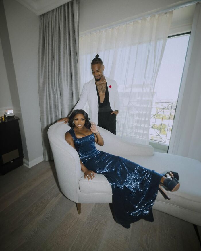 Simone Biles in a sparkling blue gown seated on a chaise lounge, with a man standing behind her in a white jacket indoors. Simone Biles in a sparkling blue gown seated on a chaise lounge, with a man standing behind her in a white jacket indoors.