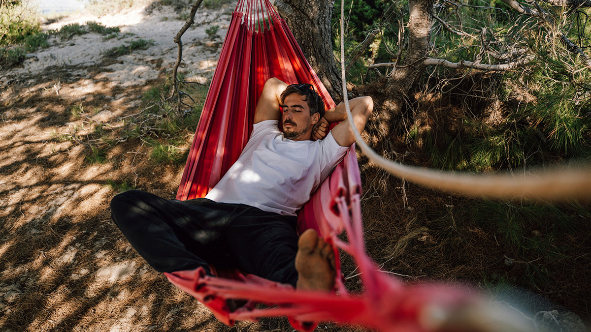 Man relaxing in a red hammock under trees, enjoying simple living outdoors with peace and minimal distractions.