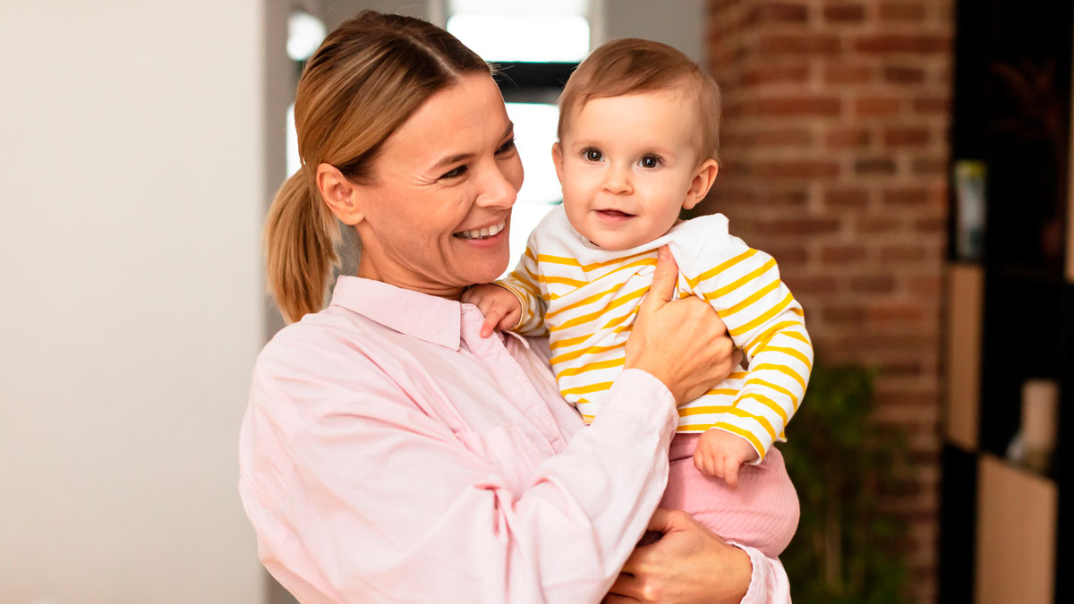 Woman smiling and holding a baby indoors, illustrating a live-in nanny job offer with difficult demands.