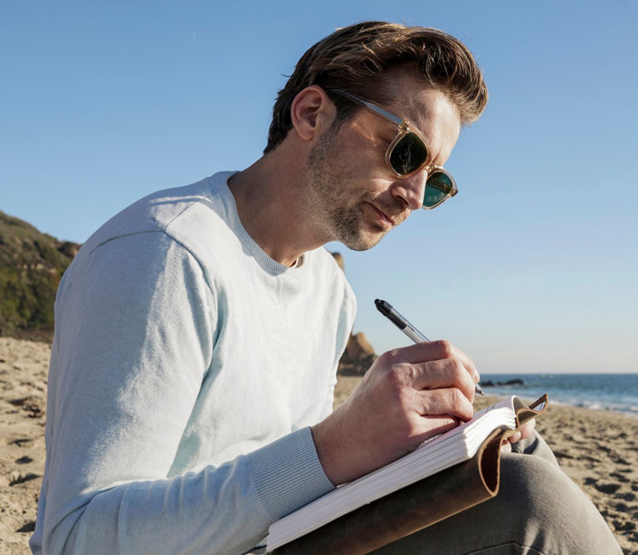 Man writing in a journal on the beach, reflecting on thoughts about marrying the wrong sister and family reactions.