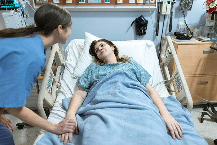 Patient lying in hospital bed covered with blanket while nurse holds her hand offering support during hospital stay.