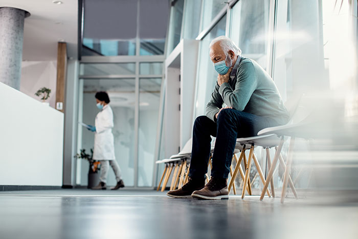 Elderly man wearing mask sitting in hospital waiting area, reflecting on unexpected surgery after small health issue.