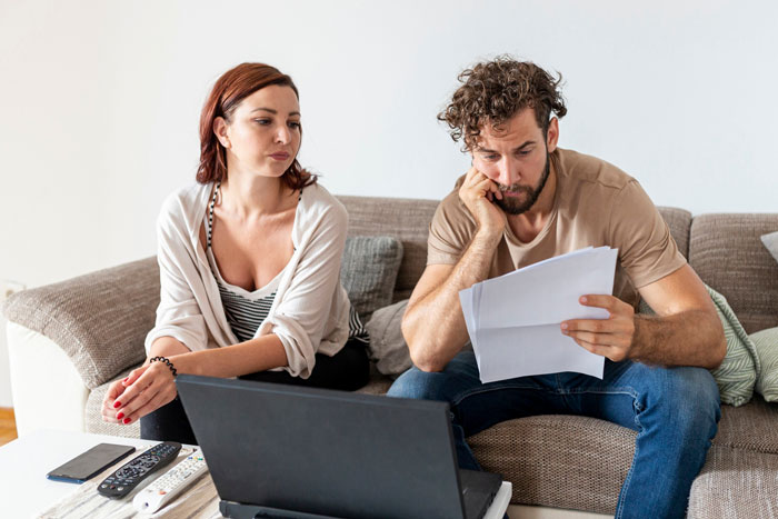 Young man mooching off parents looks worried while woman watches, both sitting on couch with laptop and papers. Young man mooching off parents looks worried while woman watches, both sitting on couch with laptop and papers.