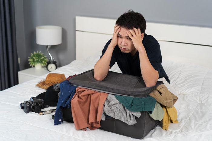 Young man looks stressed sitting on bed with overstuffed suitcase after parents kick him out for stealing money. Young man looks stressed sitting on bed with overstuffed suitcase after parents kick him out for stealing money.