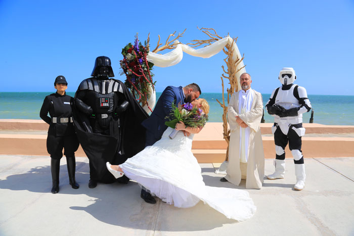 Bride and groom kiss at outdoor wedding with Star Wars characters in costume by ocean under wooden arch. Bride and groom kiss at outdoor wedding with Star Wars characters in costume by ocean under wooden arch.