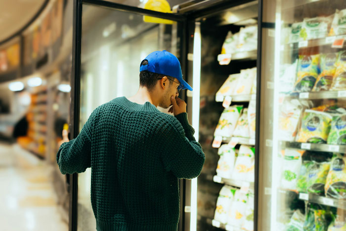 Man in blue cap and green sweater browsing frozen food aisle, reflecting on convenience store mistake and owed amount.