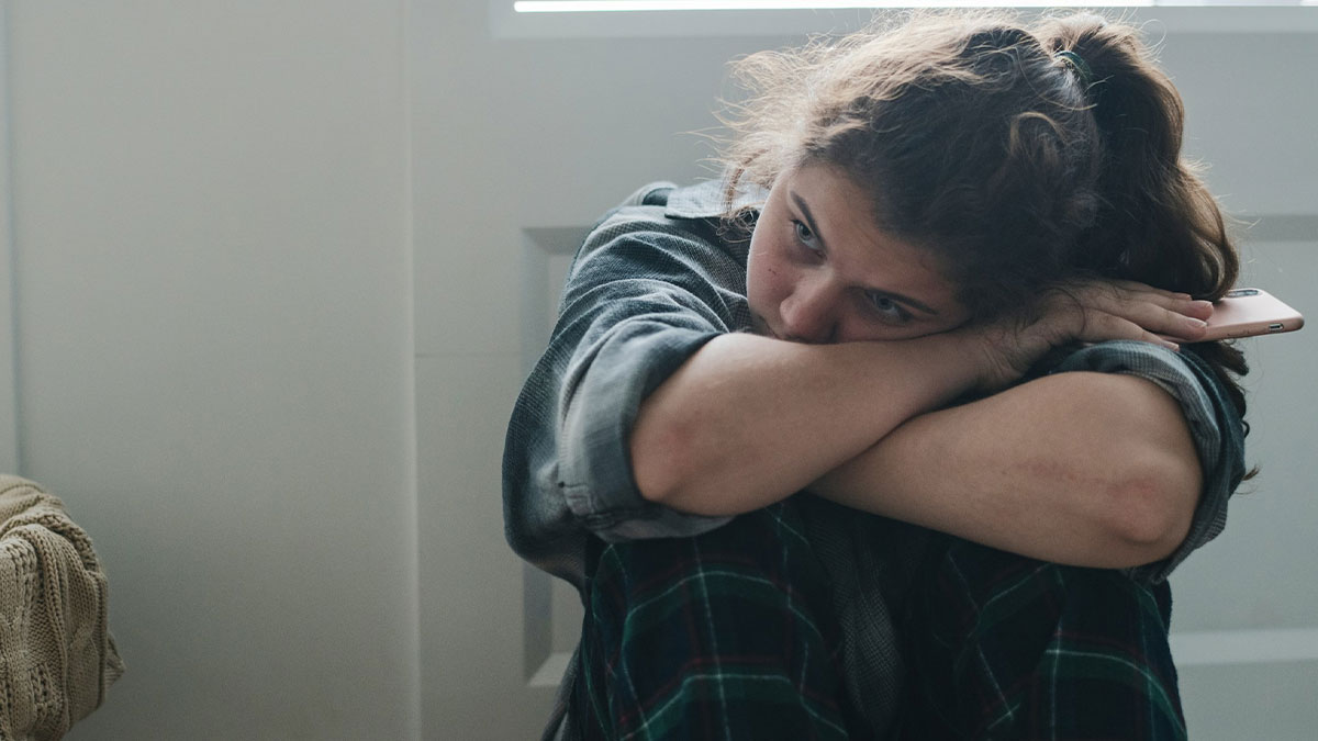Teen girl in plaid pants sitting alone, reflecting on stepmother control in family relationship, holding a phone quietly.