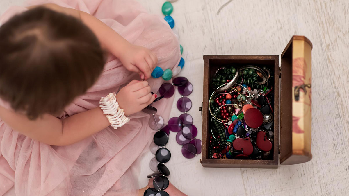 Child wearing a pink dress playing with colorful jewelry near a wooden box filled with assorted heirloom jewelry pieces