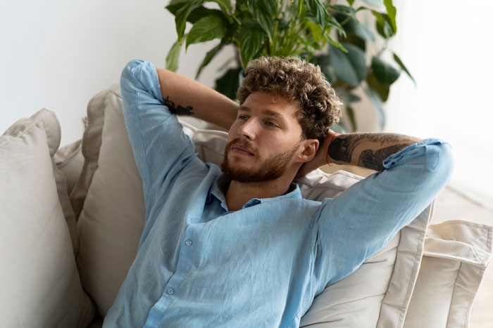 Young man with tattoos wearing a blue shirt, relaxing on a couch, representing burnout in a toxic work environment. Young man with tattoos wearing a blue shirt, relaxing on a couch, representing burnout in a toxic work environment.