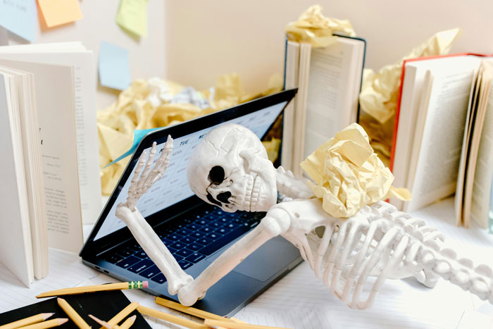 Skeleton lying over laptop surrounded by crumpled paper illustrating toxic bosses and hostile work environment burnout Skeleton lying over laptop surrounded by crumpled paper illustrating toxic bosses and hostile work environment burnout