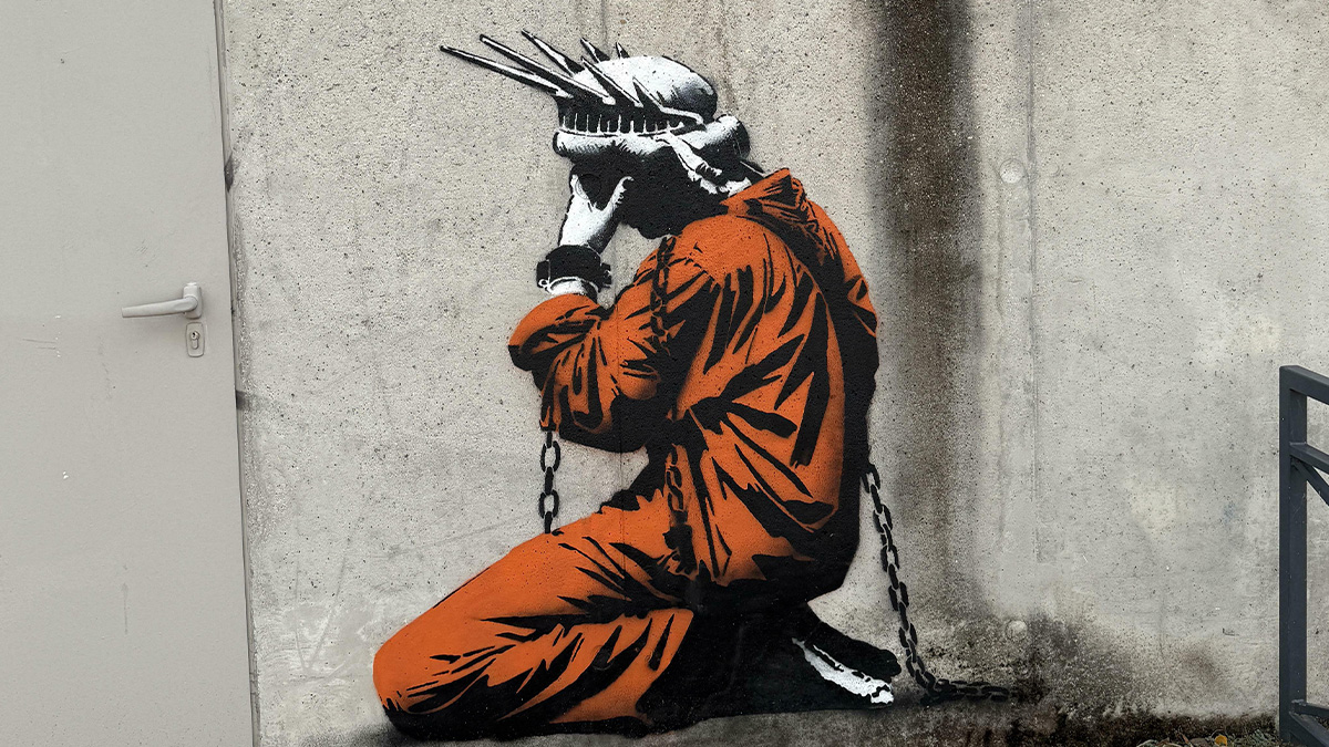 Street art of a kneeling figure in orange prison clothes wearing a crown of thorns, painted on a concrete wall with chains.