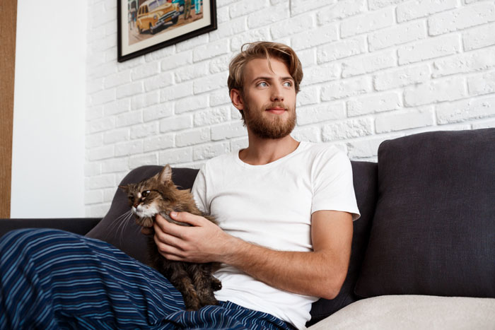 Man sitting on a couch stroking a cat during intimacy, wearing a white t-shirt and striped pants in a cozy room. Man sitting on a couch stroking a cat during intimacy, wearing a white t-shirt and striped pants in a cozy room.