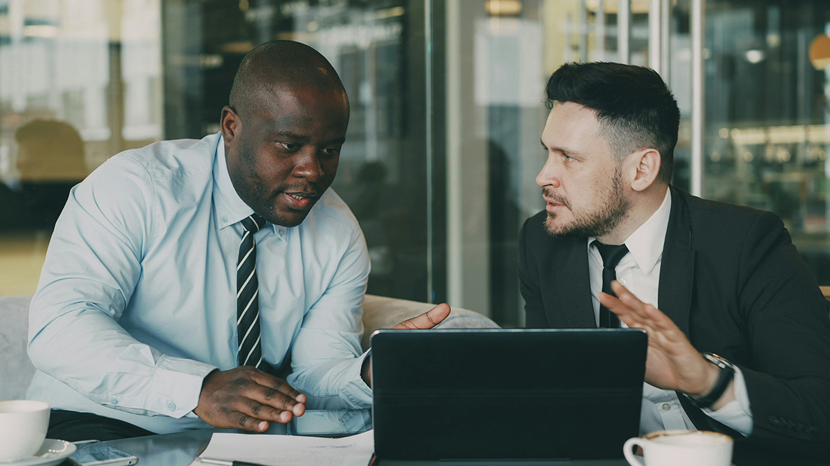 Two men in business attire discussing cancer symptoms while looking at a laptop in a modern office setting.