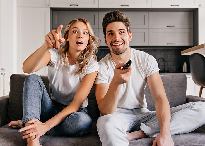 Couple sitting on couch watching TV, woman trying to teach empathy while man holds remote control and smiles. Couple sitting on couch watching TV, woman trying to teach empathy while man holds remote control and smiles.
