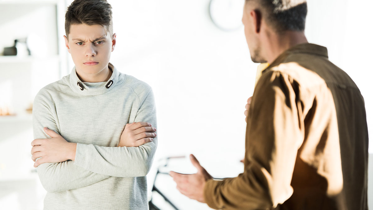 Teen boy with arms crossed looking angry while talking to an adult male, highlighting care for disabled son challenges.