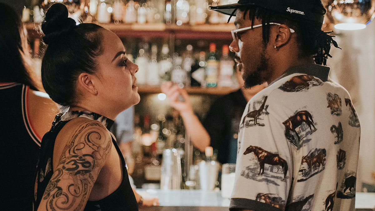 Woman and man engaged in intense conversation at a bar, hinting at exposing best friendu2019s affair to her boyfriend.