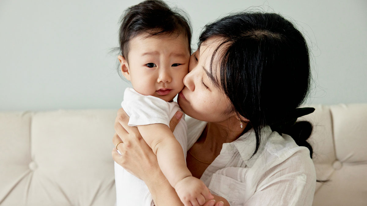 Asian mother holding and kissing toddler in a home setting, reflecting concerns about vaccinated and unvaccinated children.
