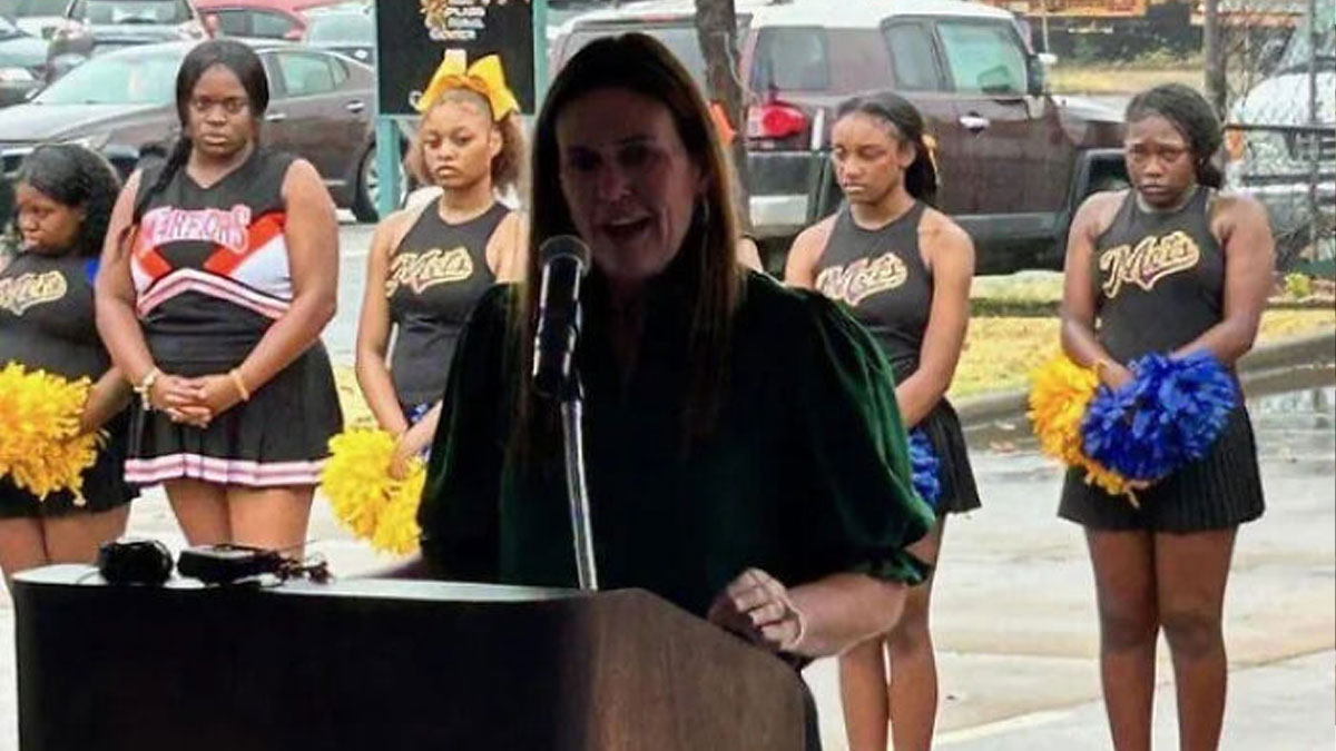 Woman speaking at a podium outdoors with cheerleaders standing behind her holding yellow and blue pom-poms.