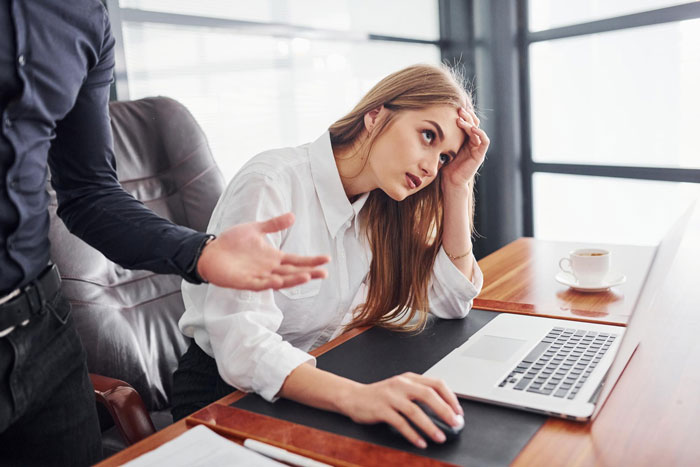 Frustrated woman at desk with laptop looking stressed while coworker gestures, illustrating work life made insufferable. Frustrated woman at desk with laptop looking stressed while coworker gestures, illustrating work life made insufferable.