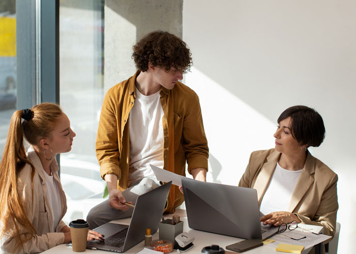 Three coworkers in a meeting, one appearing to push beliefs while others look uncomfortable at a modern office table. Three coworkers in a meeting, one appearing to push beliefs while others look uncomfortable at a modern office table.