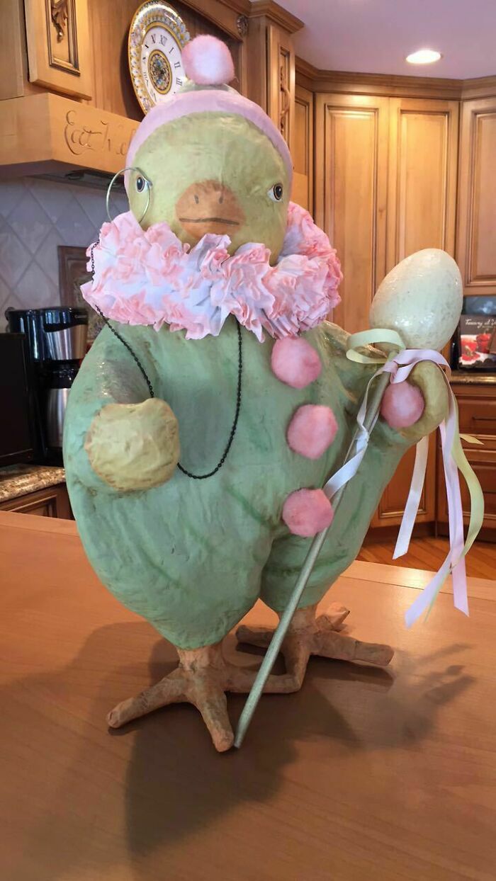 Large quirky bird figurine dressed in pastel pink ruffles and pom-poms, holding a decorated staff in a home kitchen setting.