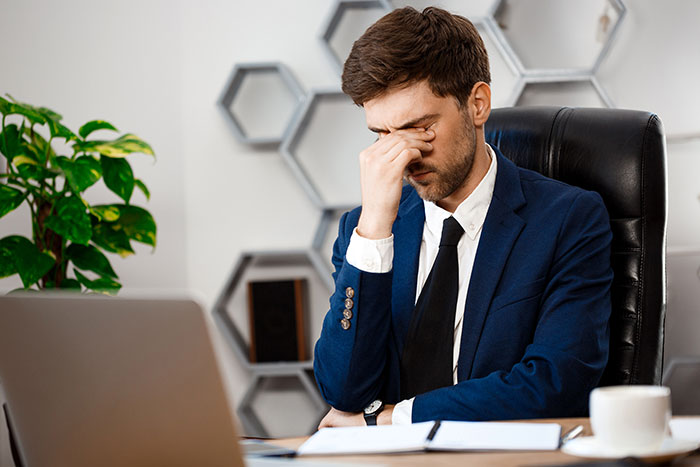 Man shuts down younger coworker flirt by looking stressed and rubbing his eyes while sitting at his office desk.